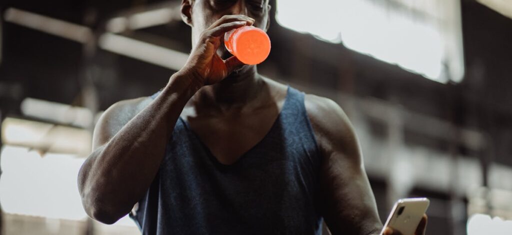 African American man drinking a sports drink while checking his smartphone in a gym setting.
