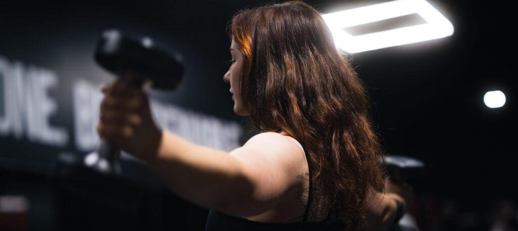 Side view of a woman exercising with dumbbells in a gym under bright lighting.