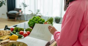 A woman in a kitchen flipping through a cookbook surrounded by fresh vegetables.